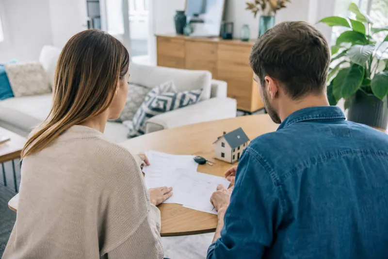 Couple comparant des documents d’assurance avec clés de voiture et maquette de maison.