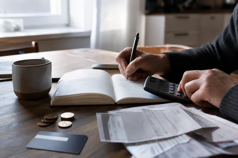 Main calculant un budget familial parmi factures, carnet et calculatrice sur une table.