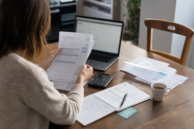 Personne comparant des relevés bancaires et calculant ses frais sur une table.