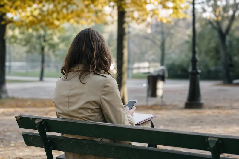 Personne assise de dos sur un banc de parc, téléphone et dossier de papiers en main